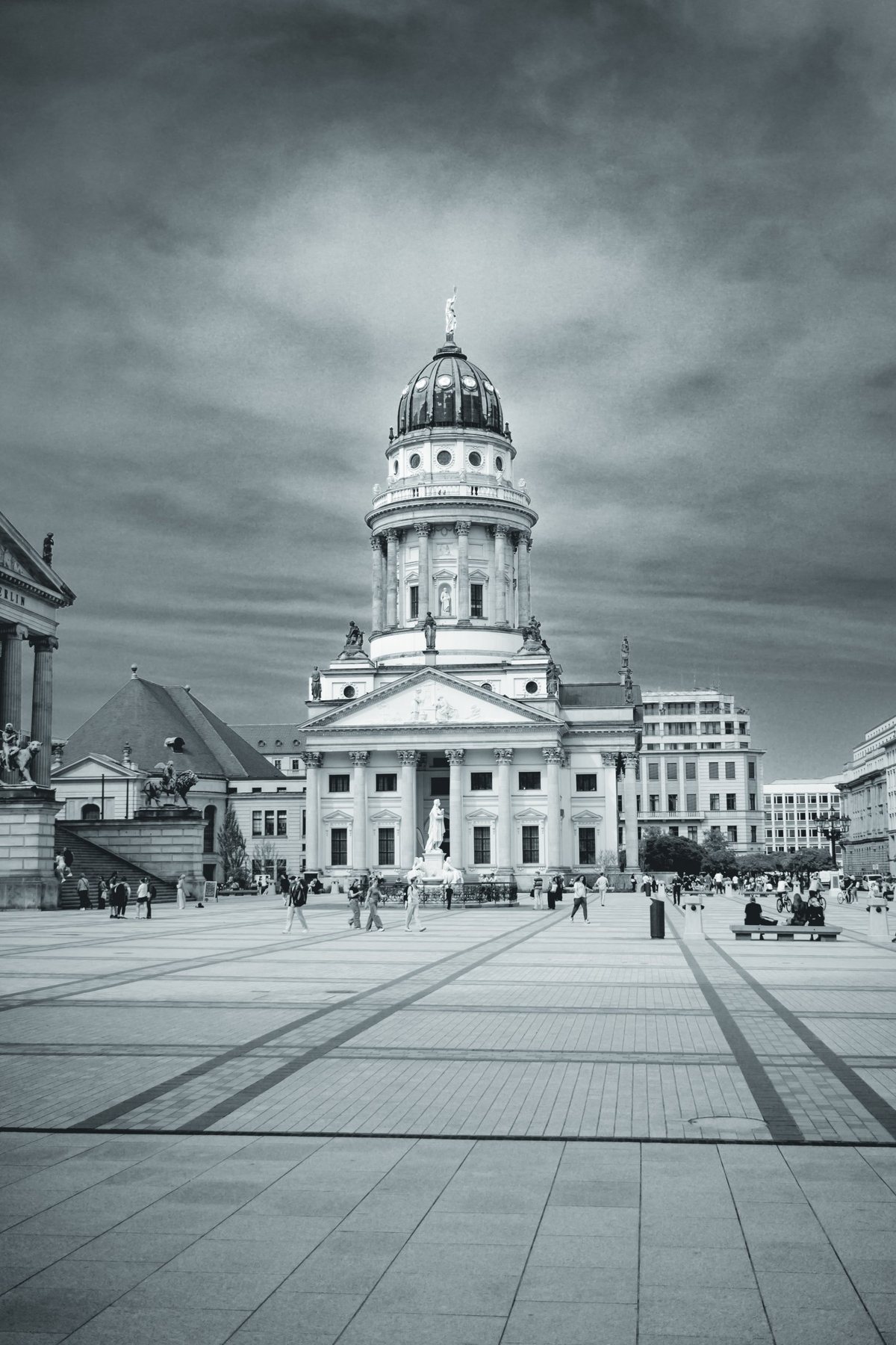 El empedrado de Gendarmenmarkt guía la mirada hasta la cúpula, con la gente pequeña bajo un cielo pesado.