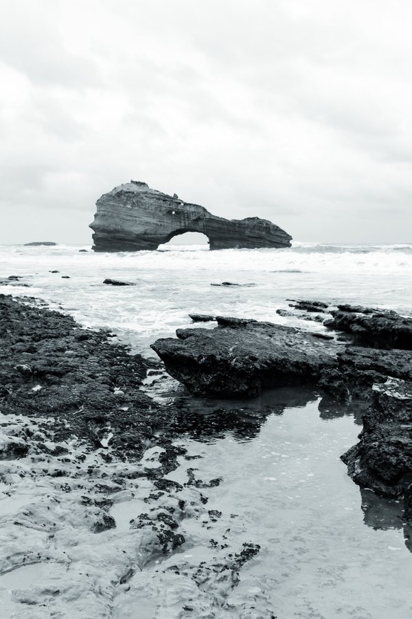 Una poza entre las rocas negras refleja el cielo; al fondo, el arco de piedra.