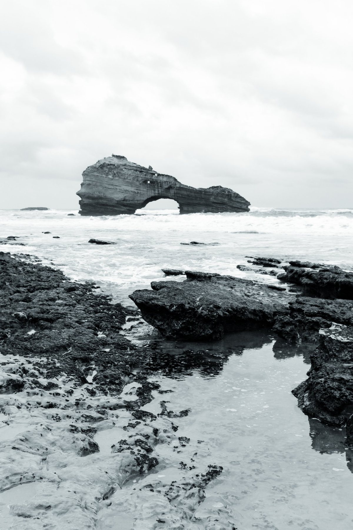 Una poza entre las rocas negras refleja el cielo; al fondo, el arco de piedra.