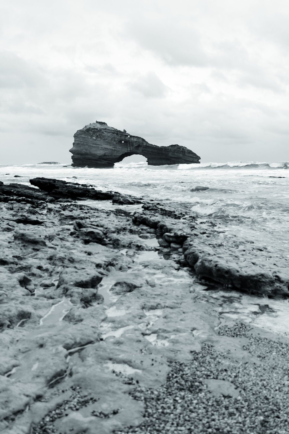 Las rocas de la marea baja conducen la mirada hasta el arco, bajo un cielo cerrado.